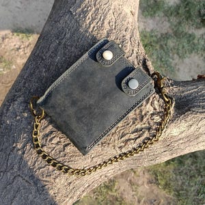 May include: A dark gray leather wallet with a chain. The wallet has two snap closures with white and silver buttons. The chain is a gold color. The wallet is sitting on a tree branch.