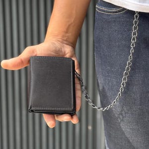 May include: A black leather wallet with a chain attached. The wallet is held in a hand, showcasing its compact, tri-fold design. The chain is silver and connects to the wallet, adding a touch of style. The wallet appears to be new and unused.