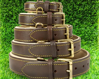 May include: A stack of five brown leather dog collars with gold-colored buckles and D-rings. Each collar is stitched with yellow thread. The collars are arranged from smallest to largest, with a green grass background.