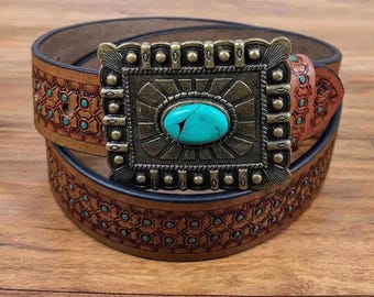 May include: A tan leather belt featuring an intricate embossed geometric pattern with small turquoise-colored inlays. The belt includes a large, rectangular, antique brass-toned buckle centered with a single, oval-shaped turquoise stone.