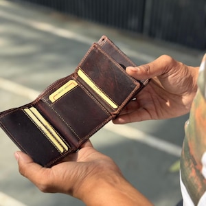 May include: A brown leather wallet, open to display credit card slots. The wallet is a tri-fold design with visible stitching and a worn, textured appearance. The interior features yellow credit cards.