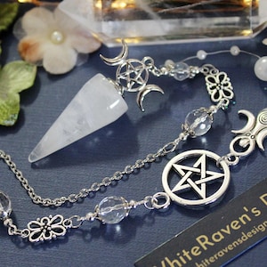 May include: A white quartz crystal pendulum with a silver chain and charms. The charms include a pentagram, a triple moon symbol, and a flower. The pendulum is hanging from a silver chain with clear crystal beads. The text "WhiteRaven's Designs" is visible in the image.