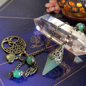 May include: A green crystal pendulum with a bronze chain and ornate metal accents hangs over a clear quartz crystal. The pendulum is adorned with two green beads and a charm shaped like a tree of life.