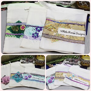 May include: Five white fabric pouches with different embellishments. Each pouch has a label that says "White Raven Designs".