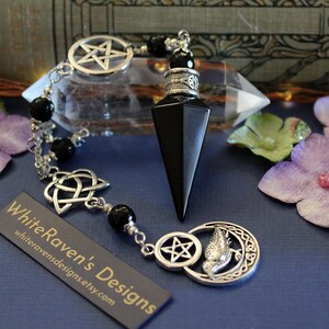 K&ouml;nnte beinhalten: Ein schwarzes Obsidian-Pendel mit einer silbernen Kette und Anh&auml;ngern, darunter ein Pentagramm, ein keltischer Knoten und ein Mond mit Gesicht. Das Pendel hat eine Spitze und wird auf einer blauen Oberfl&auml;che pr&auml;sentiert.