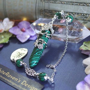 May include: A green and black malachite pendant with silver floral detailing and a chain. The pendant is cylindrical with a patterned design. The chain features decorative beads and a leaf-shaped charm.