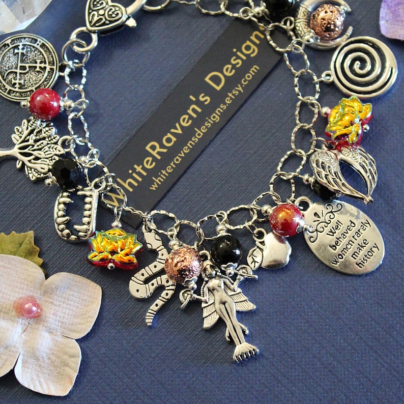 May include: A silver-toned charm bracelet featuring various pendants including a tree, a snake, a fairy, vampire fangs, and an oval tag with the text 'Well behaved women rarely make history'. The bracelet includes red, black, and metallic beads.