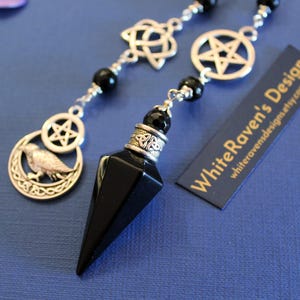 K&ouml;nnte beinhalten: Ein schwarzes, facettiertes Pendel mit silbernen Akzenten und einem keltischen Knotenmuster, das an einer Perlenkette mit silbernen Anh&auml;ngern h&auml;ngt. Zu den Anh&auml;ngern geh&ouml;ren ein Pentagramm, ein keltischer Knoten und ein Vogel in einem Halbmond. Der Text "White Raven's Design" ist sichtbar.