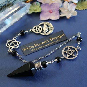 K&ouml;nnte beinhalten: Ein schwarzes Pendel mit einer silbernen Kette und dekorativen Anh&auml;ngern, darunter ein Pentagramm, ein Mond und ein Knotendesign. Das Pendel hat eine Spitze und ist mit schwarzen Perlen verziert. Der Text "WhiteRaven's Designs" ist sichtbar.