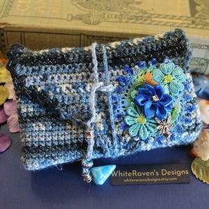 May include: A rectangular crochet pouch in shades of blue, black, and white with metallic thread. It features a blue ribbon flower, teal lace flowers, a small metal fairy charm, and a dangling blue stone bead. Text reads WhiteRaven's Designs.