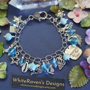 May include: Silver-toned charm bracelet with blue beads and sea-themed charms, including a starfish, seashell, and mermaid. The bracelet has a lobster clasp and a tag that reads "Dreaming of the Sea". The bracelet is displayed on a dark blue surface.