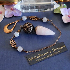 May include: A pendulum featuring a pointed rose quartz stone, a dark metal filigree sphere, and light-colored beads on a copper-toned chain. A copper teardrop charm is attached. A black rectangular tag displays the text WhiteRaven's Designs.