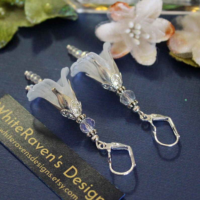 May include: A pair of silver earrings with white flower-shaped charms and clear crystal beads. The earrings have a hook closure.