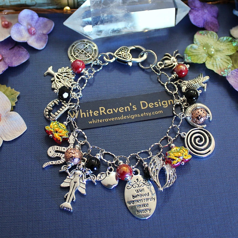 May include: A silver-toned charm bracelet featuring various pendants including a mermaid, a tree, a spiral, wings, and a tag inscribed with the text Well behaved women rarely make history. The bracelet includes red and black beads and a heart-shaped clasp.