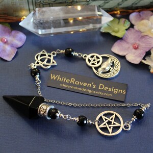 K&ouml;nnte beinhalten: Ein schwarzes und silbernes Pendel mit einem spitz zulaufenden Onyx-Kristall und einer Kette mit schwarzen Perlen. Die Kette hat silberne Anh&auml;nger, darunter ein Pentagramm, einen keltischen Knoten und einen Mond mit einem Vogel. Die Visitenkarte lautet "WhiteRaven's Designs".