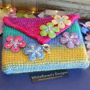 May include: A handmade, rainbow-colored crochet clutch with a flap closure. The clutch is adorned with five fabric flower appliqués in pink, blue, green, yellow, and purple, each embellished with sequins. A small silver-toned ring clasp secures the flap. The text "WhiteRaven's Designs" is visible.
