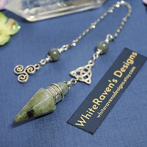May include: A green stone pendulum with a silver-toned metal cap, attached to a silver chain featuring two grey beads, a silver triskele charm, and a silver triquetra charm. A black rectangular tag displays the text WhiteRaven's Designs.