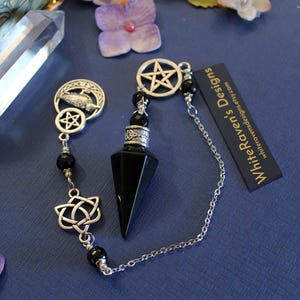 K&ouml;nnte beinhalten: Ein schwarzes Obsidian-Pendel mit silberfarbenen Akzenten, darunter ein Pentagramm, ein keltischer Knoten und eine Kette. Das Pendel ist f&uuml;r Wahrsagung und spirituelle Praktiken konzipiert. Der Hintergrund ist eine dunkelblaue Oberfl&auml;che mit dekorativen Elementen.