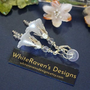May include: A pair of silver earrings with white flower-shaped charms and crystal beads. The earrings have a hook closure. The earrings are on a blue background with a business card that says "WhiteRaven's Designs" and a website address.