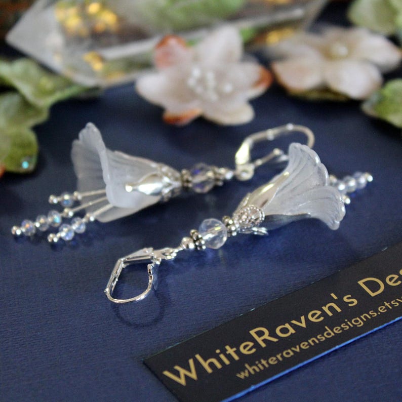 May include: Silver earrings with a white flower design and crystal accents. The earrings are dangle style and have a hook closure.