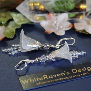 May include: A pair of silver earrings with white flower-shaped charms and clear crystal beads. The earrings are hanging from silver hooks.