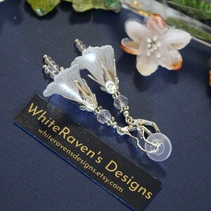 May include: A pair of silver earrings with white flower-shaped charms and crystal beads. The earrings are attached to a silver hook with a clear rubber backing. The earrings are displayed on a black card with the text "White Raven's Designs whiteravensdesigns.etsy.com".