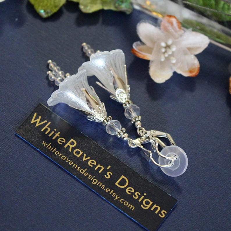 May include: A pair of silver earrings with white flower-shaped charms and crystal beads. The earrings are attached to a silver hook with a clear rubber backing. The earrings are displayed on a black card with the text "White Raven's Designs whiteravensdesigns.etsy.com".