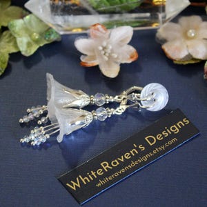 May include: White flower-shaped earrings with crystal beads and silver accents. The earrings are clip-on style. The earrings are displayed on a blue surface with a business card that reads "White Raven's Designs" and a website address.