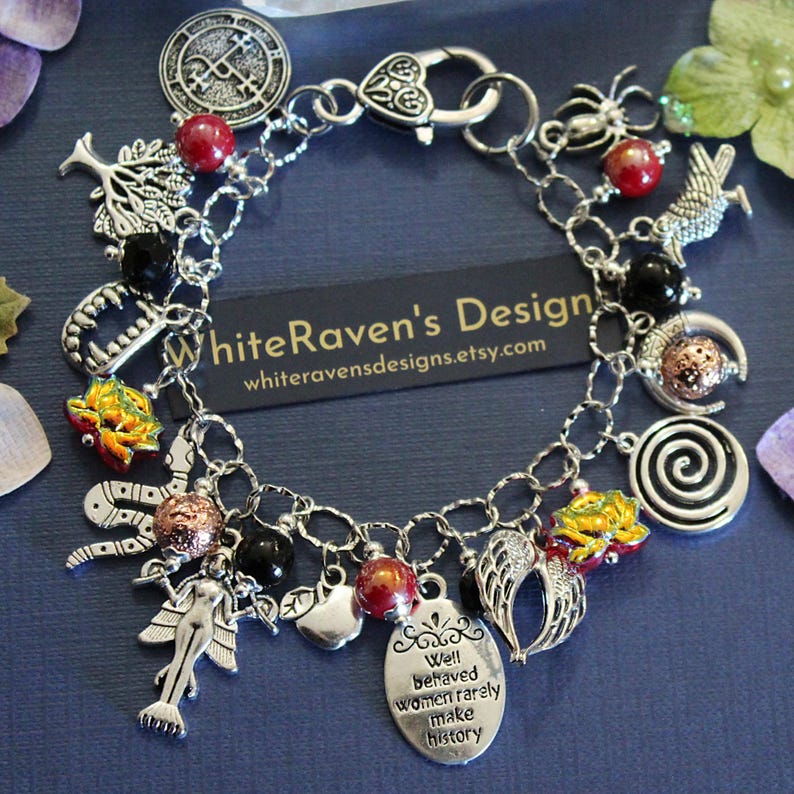 May include: A silver-tone charm bracelet featuring various pendants including a tree, a spider, a snake, wings, a mermaid, and an oval tag with the text "Well behaved women rarely make history." The bracelet includes red, black, and textured copper beads.