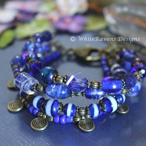 a close up photo of a multistrand bracelet with dark blue beads with a white stripe. the metal accents and spiral charms are a bronze colour.