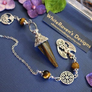 May include: A silver-toned pendulum with a tiger's eye point and decorative accents. The pendulum is connected to a chain with tiger's eye beads and ornate silver charms. The image includes a business card that reads "WhiteRaven's Designs".