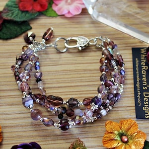 May include: A multi-strand bracelet featuring various shades of purple and clear faceted glass beads, interspersed with small silver-toned metal spacers. The bracelet includes a silver-toned heart-shaped lobster clasp and a dangling bead charm.
