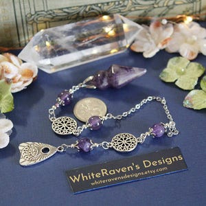 May include: Amethyst pendulum with a silver chain and decorative charms. The pendulum is hanging from a silver chain with amethyst beads and two silver charms. The charms are in the shape of a circle with a floral design and a triangle with an eye design. The pendulum is on a blue surface with a white card that says "WhiteRaven's Designs" and a website address.