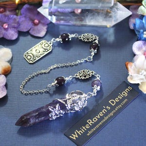 May include: A silver pendulum with a purple amethyst crystal point. The chain has decorative beads and a sun and moon charm. The product tag reads "WhiteRaven's Designs". The website address is also visible.