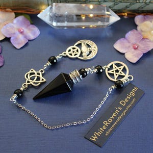 K&ouml;nnte beinhalten: Ein schwarzes Obsidian-Pendel mit silberfarbenen Akzenten und einer Kette. Das Pendel hat eine Spitze und ist mit schwarzen Perlen und dekorativen Anh&auml;ngern verziert, darunter ein Halbmond, ein Pentagramm und ein keltischer Knoten. Das Etikett lautet "WhiteRavenDesigns.etsy.com".