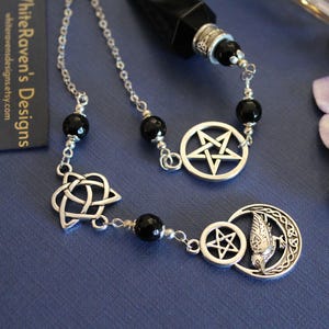 K&ouml;nnte beinhalten: Eine silberfarbene Halskette mit schwarzen Perlen und verschiedenen Anh&auml;ngern. Zu den Anh&auml;ngern geh&ouml;ren ein Pentagramm, ein keltischer Knoten und ein Halbmond mit einem Vogel. Die Halskette wird auf einer blauen Oberfl&auml;che pr&auml;sentiert. Der Text "WhiteRaven's Designs" ist sichtbar.