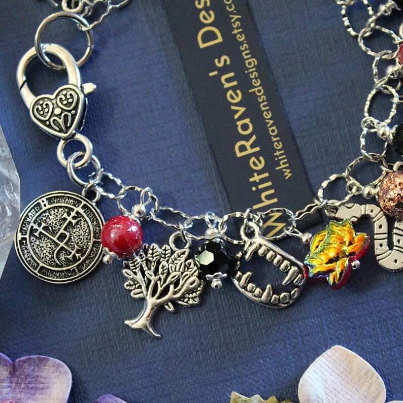 May include: A silver-toned charm bracelet featuring a heart-shaped lobster clasp and various charms including a circular sigil, a red bead, a tree, a black bead, vampire fangs, a colorful flower, and a snake, with a tag reading WhiteRaven's Designs.