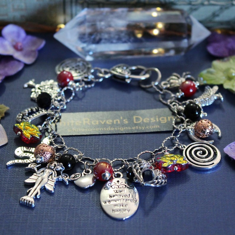 May include: A silver-toned charm bracelet featuring various metal charms, including a figure with wings, a spiral, and an oval pendant inscribed with the text "Well behaved women rarely make history." It includes red, black, and textured copper-colored beads.