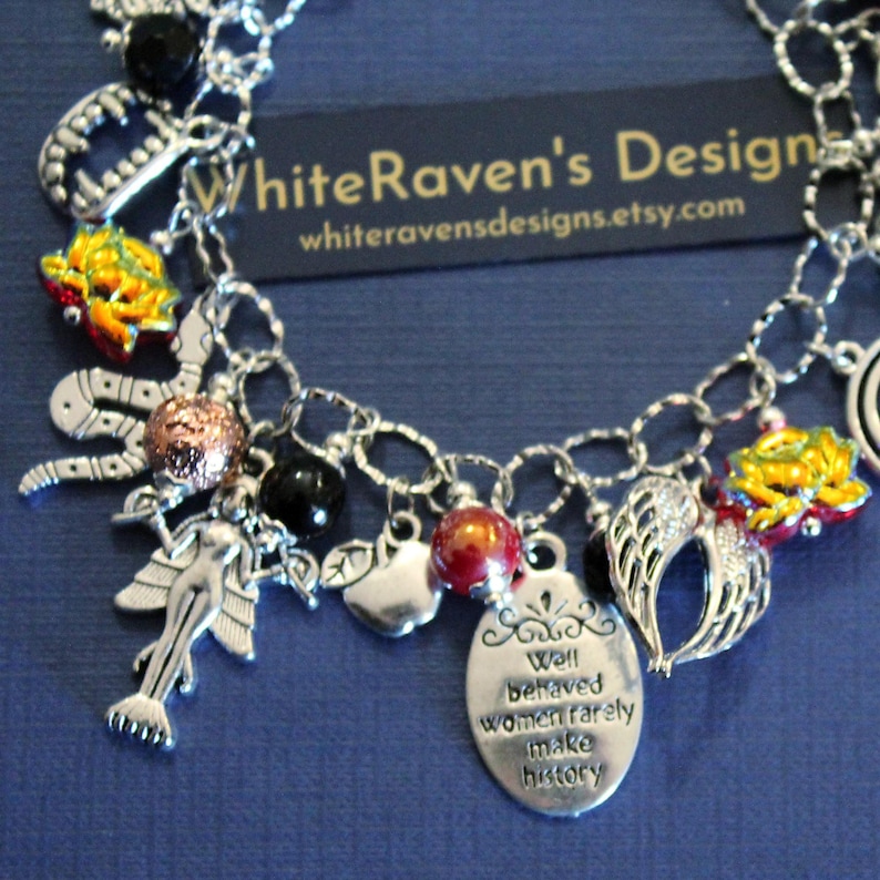 May include: A silver-toned charm bracelet featuring various pendants including a mermaid, angel wings, an apple, a snake, and a red and gold bead. An oval charm is engraved with the text, Well behaved women rarely make history.