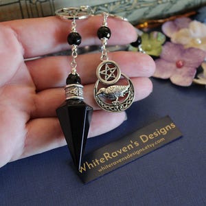K&ouml;nnte beinhalten: Ein schwarzes Onyx-Pendel mit einer silberfarbenen Kette, Perlen und Anh&auml;ngern. Ein Anh&auml;nger zeigt einen Raben in einem Halbmond und einem Pentagramm. Der andere Anh&auml;nger ist ein Pentagramm. Das Etikett lautet "WhiteRaven's Designs".