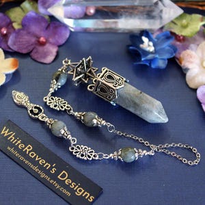 May include: A silver pendulum with a grey crystal and decorative chain. The pendulum has a star-shaped top and small, round grey beads. The image includes the text "WhiteRaven's Designs" on a black tag.