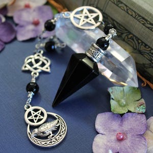 K&ouml;nnte beinhalten: Ein schwarzes Kristallpendel mit silberfarbenen Akzenten, darunter ein Pentagramm, ein keltischer Knoten und ein Rabenmotiv. Das Pendel h&auml;ngt an einer Kette mit schwarzen Perlen. Ein klarer Quarz-Kristall und lila Blumen im Hintergrund.