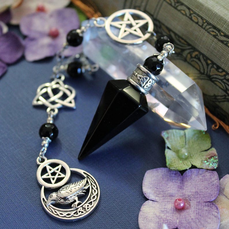 K&ouml;nnte beinhalten: Ein schwarzes Kristallpendel mit silberfarbenen Akzenten, darunter ein Pentagramm, ein keltischer Knoten und ein Rabenmotiv. Das Pendel h&auml;ngt an einer Kette mit schwarzen Perlen. Ein klarer Quarz-Kristall und lila Blumen im Hintergrund.
