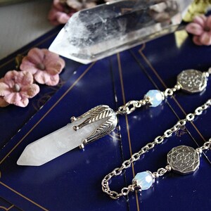 May include: A white quartz crystal pendulum with a silver chain and decorative accents. The pendulum is hanging over a blue surface with a clear quartz crystal in the background.