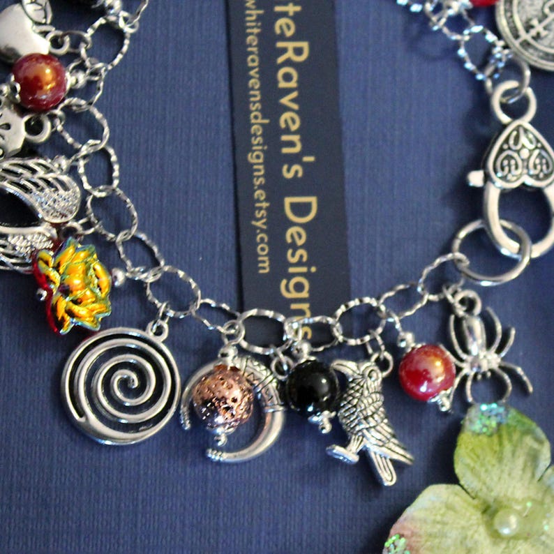 May include: A silver charm bracelet featuring various pendants including a spiral, a crescent moon with a textured sphere, a raven, a spider, a flower, and red and black beads. A heart-shaped lobster clasp and a tag reading WhiteRaven'sDesigns.etsy.com.