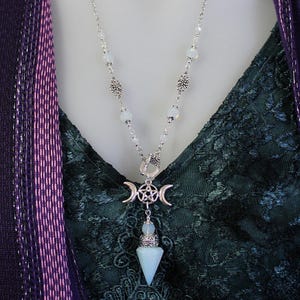 May include: A silver necklace with a white opal pendant and a triple moon charm. The pendant is a point and has a silver pentagram in the center. The necklace is on a green and white lace background.