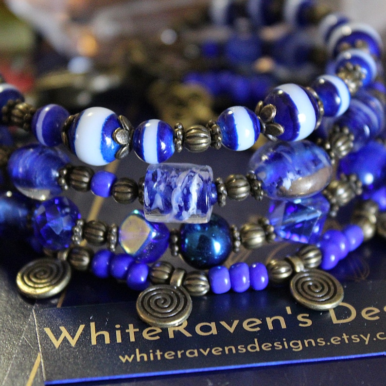 K&ouml;nnte beinhalten: Ein mehrreihiges Armband mit blauen und wei&szlig;en Glasperlen, blau-wei&szlig;en Streifenperlen und goldfarbenen Metallperlen. Das Armband enth&auml;lt au&szlig;erdem zwei goldfarbene Metallanh&auml;nger mit einem Spiraldesign. Das Armband befindet sich auf einem blauen Hintergrund mit dem Text "WhiteRaven's Designs" und "whiteravensdesigns.etsy.com".