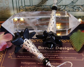 May include: Black flower dangle earrings with silver filigree accents and black beads. The earrings are displayed on a clear crystal and a book with the text "Rave Designs ensdesigns.etsy.com Whiteraven Pendulums. Prayads and witchy Jewellery Instagram: whitensdesias Facebook: whiterav designs".