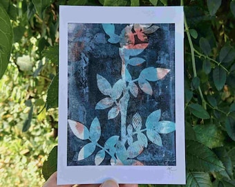 Könnte beinhalten: Ein botanischer Kunstdruck mit einer stilisierten Pflanze mit blau-pfirsichfarbenen Blättern vor einem dunkelblauen Hintergrund. Der Druck befindet sich auf weißem Karton und wird vor einem Hintergrund aus grünen Blättern gehalten.