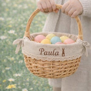 Könnte beinhalten: Ein geflochtener Osterkorb mit pastellfarbenen Eiern. Der Korb hat einen Henkel und ein Stofffutter mit dem gestickten Namen "Paula" und einer kleinen Hasen-Silhouette. Der Korb wird vor einem verschwommenen Hintergrund aus Blumen gehalten.